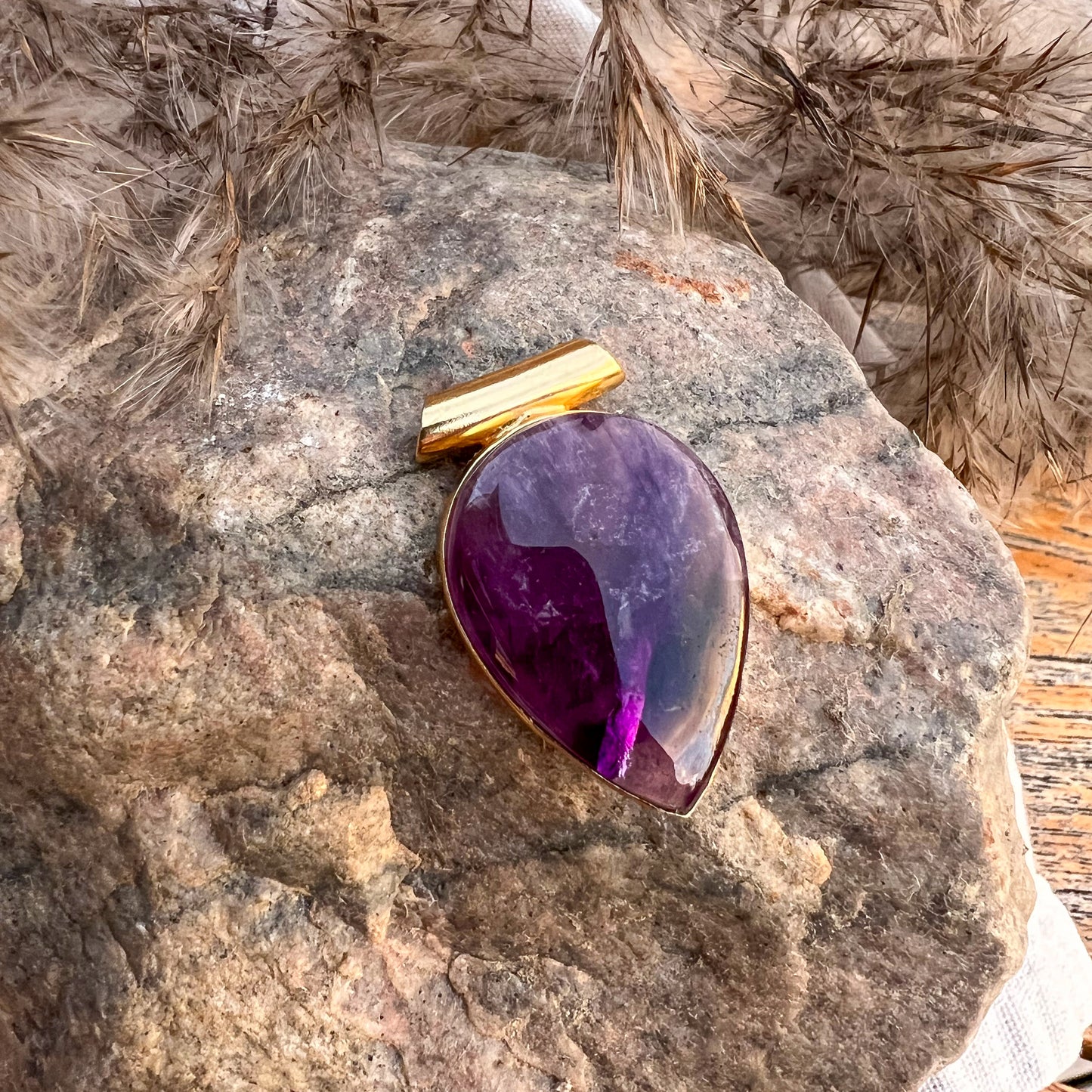 Ein tropfenförmiger Anhänger aus poliertem Amethyst, eingefasst in eine goldene Halterung, liegt auf einem groben Stein. Im Hintergrund sind getrocknete Gräser zu sehen, die eine natürliche, rustikale Atmosphäre schaffen.