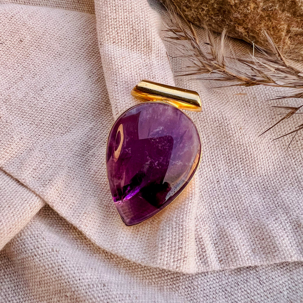 
                  
                    Ein tropfenförmiger Anhänger aus poliertem Amethyst, eingefasst in eine goldene Halterung, liegt auf einem beigen Stofftuch. Im Hintergrund sind getrocknete Gräser zu sehen, die eine natürliche und rustikale Atmosphäre schaffen.
                  
                