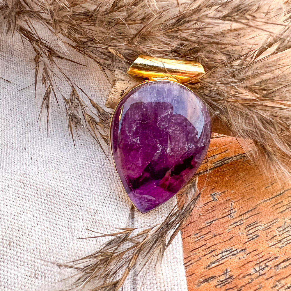 Ein tropfenförmiger Anhänger aus poliertem Amethyst, eingefasst in eine goldene Halterung, liegt auf einem beigen Stofftuch. Im Hintergrund sind getrocknete Gräser zu sehen, die eine natürliche und rustikale Atmosphäre schaffen.