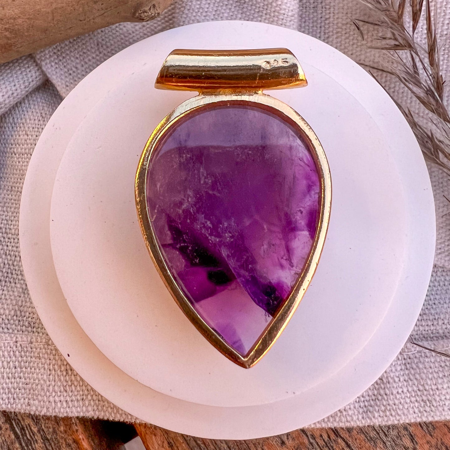 
                  
                    Ein tropfenförmiger Anhänger aus poliertem Amethyst, eingefasst in eine goldene Halterung, liegt auf einem weißen Keramikteller, der auf einem beigen Stofftuch platziert ist. Im Hintergrund sind getrocknete Gräser und ein Teil eines Holztisches zu sehen, die eine natürliche und rustikale Atmosphäre schaffen.
                  
                