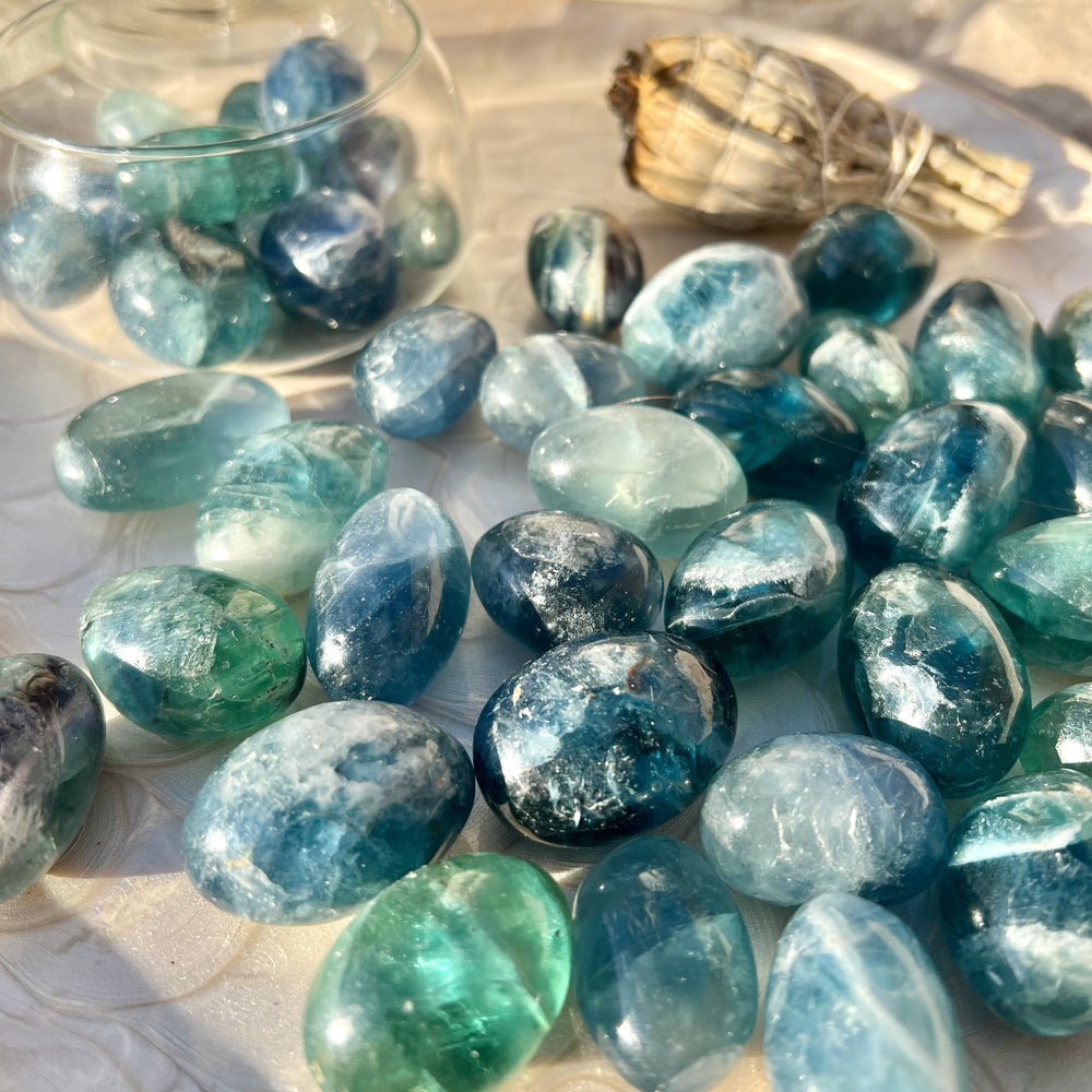 Ein Arrangement von blauen Fluorit-Trommelsteinen auf einem dekorativen Teller in warmem Sonnenlicht. Die Steine zeigen intensive Blautöne mit grünen Nuancen und natürlicher Transparenz. Im Hintergrund stehen ein Glasgefäß mit weiteren Steinen und ein Bündel weißer Salbei als Dekoration.