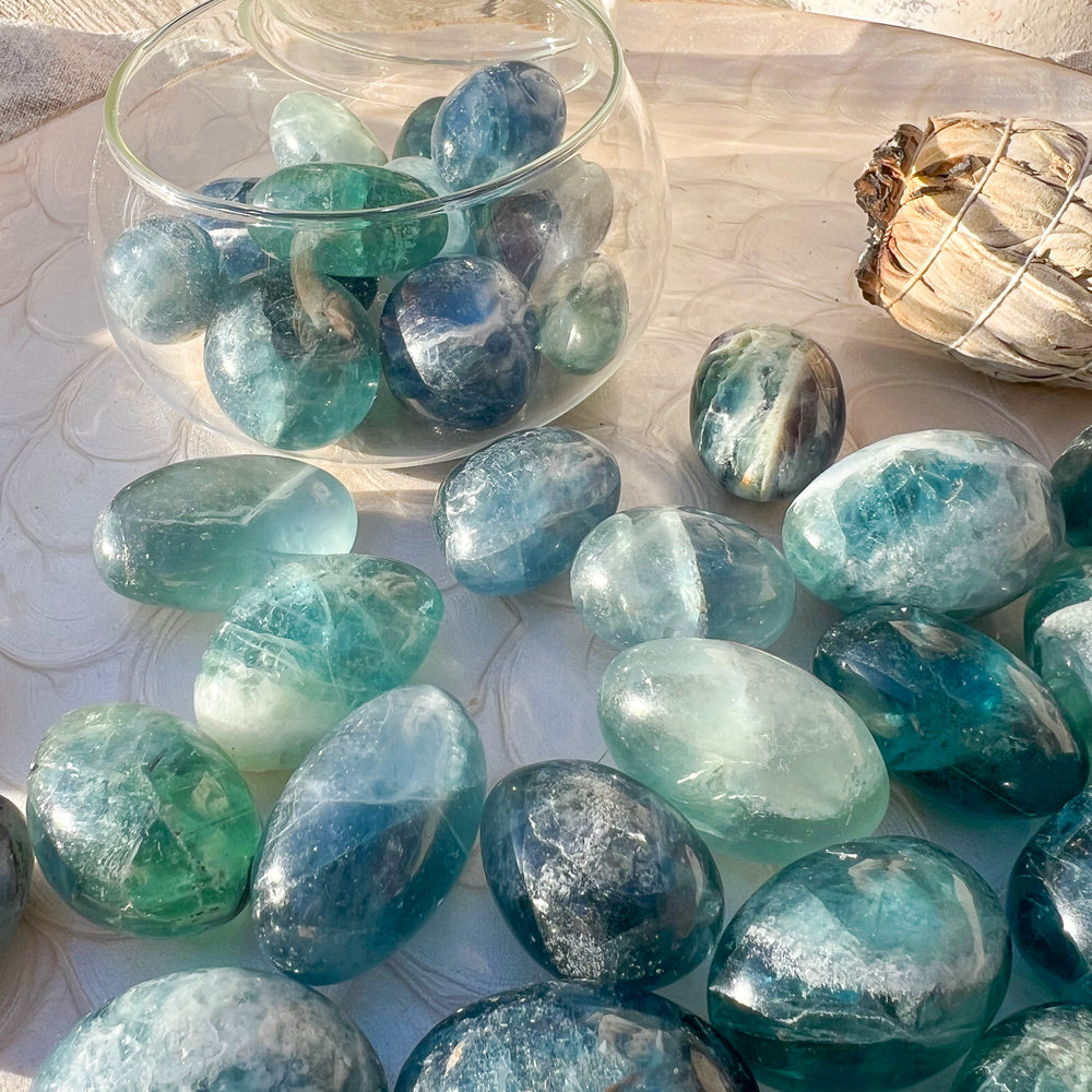 Ein Arrangement von blauen Fluorit-Trommelsteinen auf einem dekorativen Teller in warmem Sonnenlicht. Die Steine zeigen intensive Blautöne mit grünen Nuancen und natürlicher Transparenz. Im Hintergrund stehen ein Glasgefäß mit weiteren Steinen und ein Bündel weißer Salbei als Dekoration.