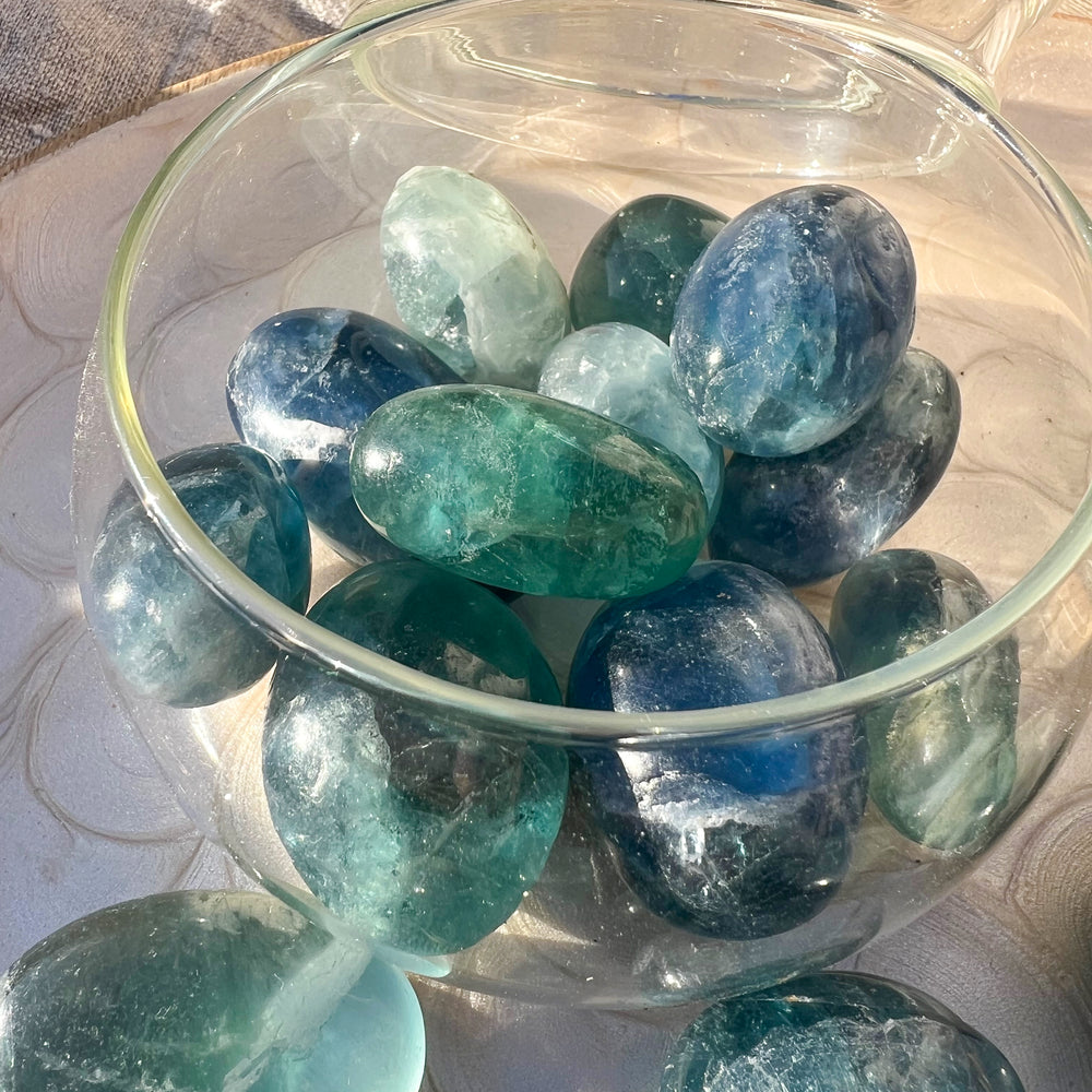 Detailaufnahme eines Glasgefäßes, das mit blauen und grünen Fluorit-Trommelsteinen gefüllt ist. Die Steine schimmern sanft im Sonnenlicht, das ihre Transparenz und natürliche Maserung hervorhebt.
