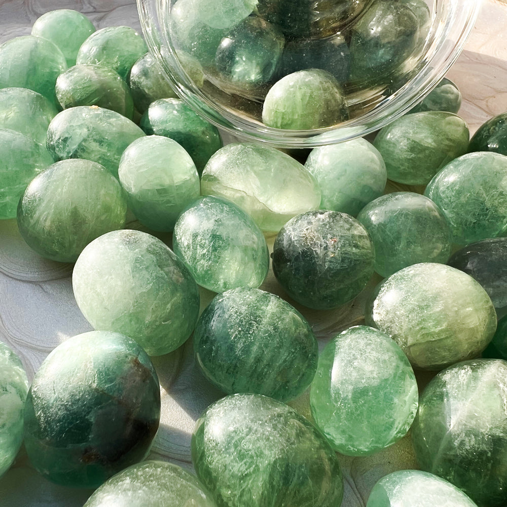 Nahaufnahme von mehreren grünen Fluorit-Trommelsteinen auf einem strukturierten Teller. Die Steine zeigen verschiedene grüne Nuancen und natürliche Einschüsse, die im Sonnenlicht schimmern.