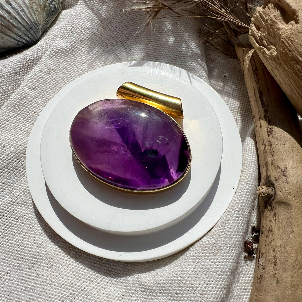 Das Bild zeigt einen ovalen Amethyst-Anhänger, der in eine vergoldete Fassung eingebettet ist. Der Edelstein schimmert in intensiven Violetttönen, die im Licht facettenreich erscheinen. Der Anhänger liegt auf einem hellen Stoff, umgeben von natürlichen Elementen wie Treibholz und einer Muschelschale, was eine harmonische und naturverbundene Atmosphäre schafft. 