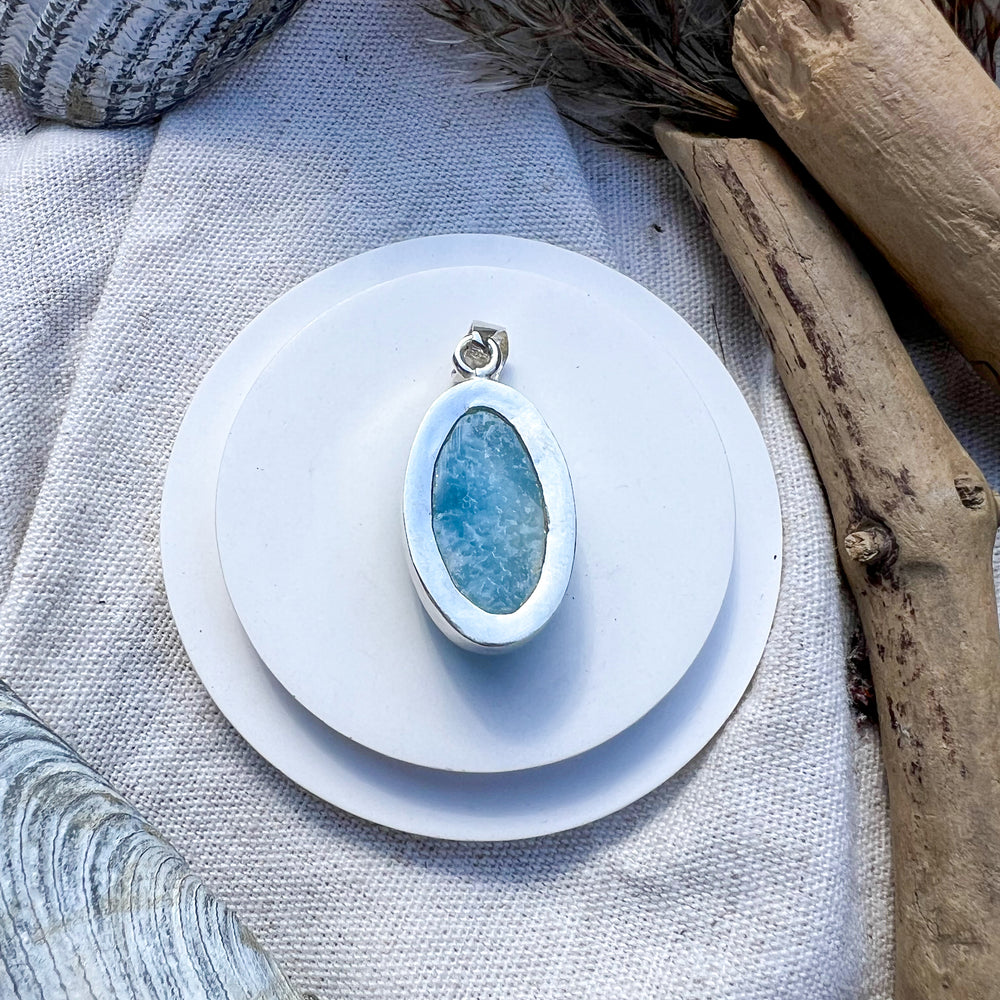 Ovaler Larimar-Anhänger in 925er Silberfassung, präsentiert auf weißem Untergrund mit Treibholz und Muscheln als dekorativem Hintergrund.