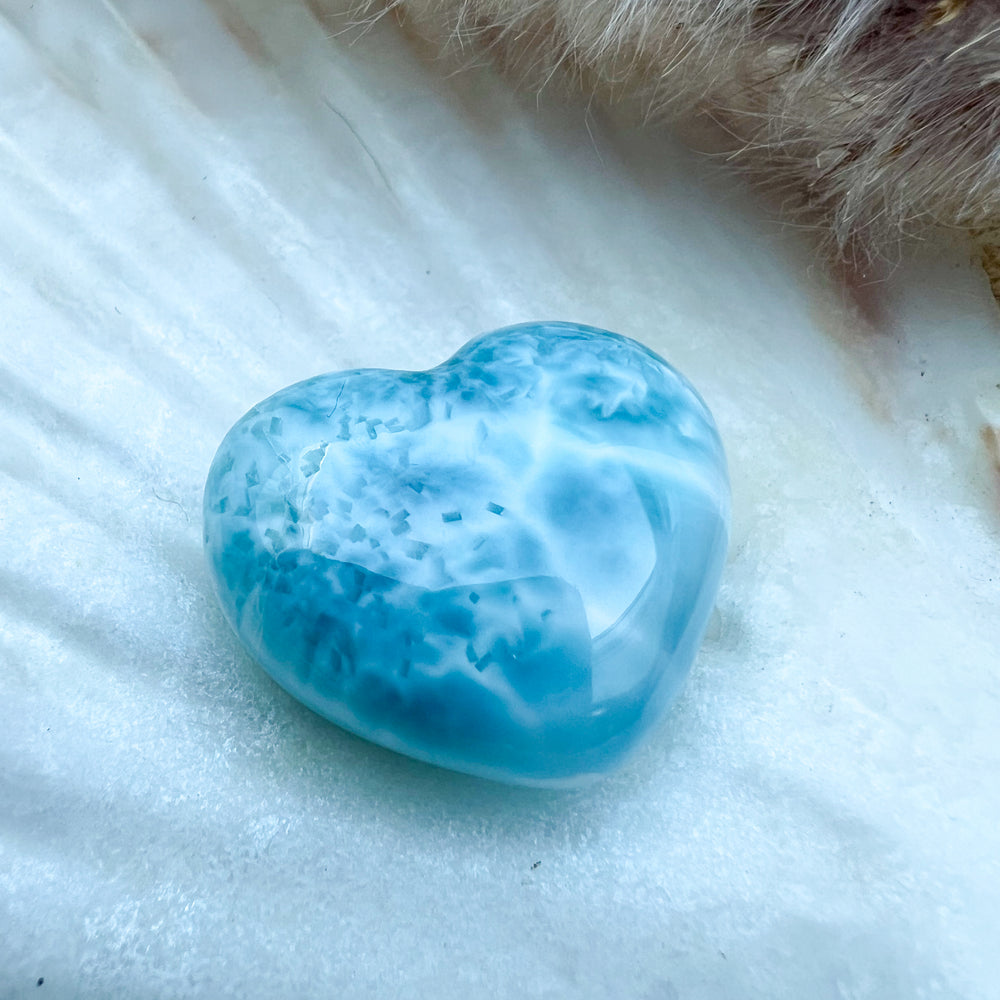 Handgearbeitetes Larimar Herz in meeresblau mit weißen Akzenten, präsentiert auf einer Muschelschale – einzigartiger Edelstein aus der Dominikanischen Republik.