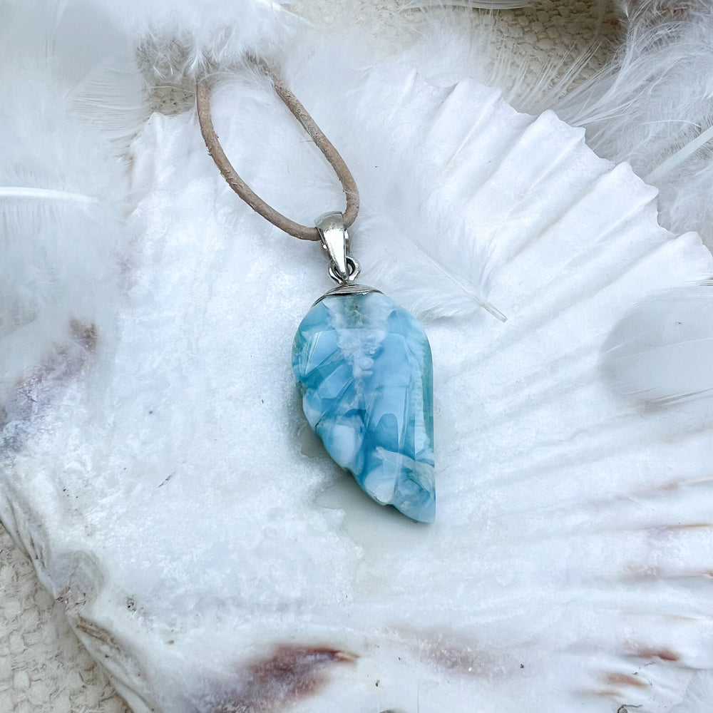 Larimar Engelsflügel-Anhänger aus 925er Sterlingsilber, präsentiert auf einer weißen Muschelschale, umgeben von Federn, mit natürlichem Lederband – ein himmlisches Schmuckstück.