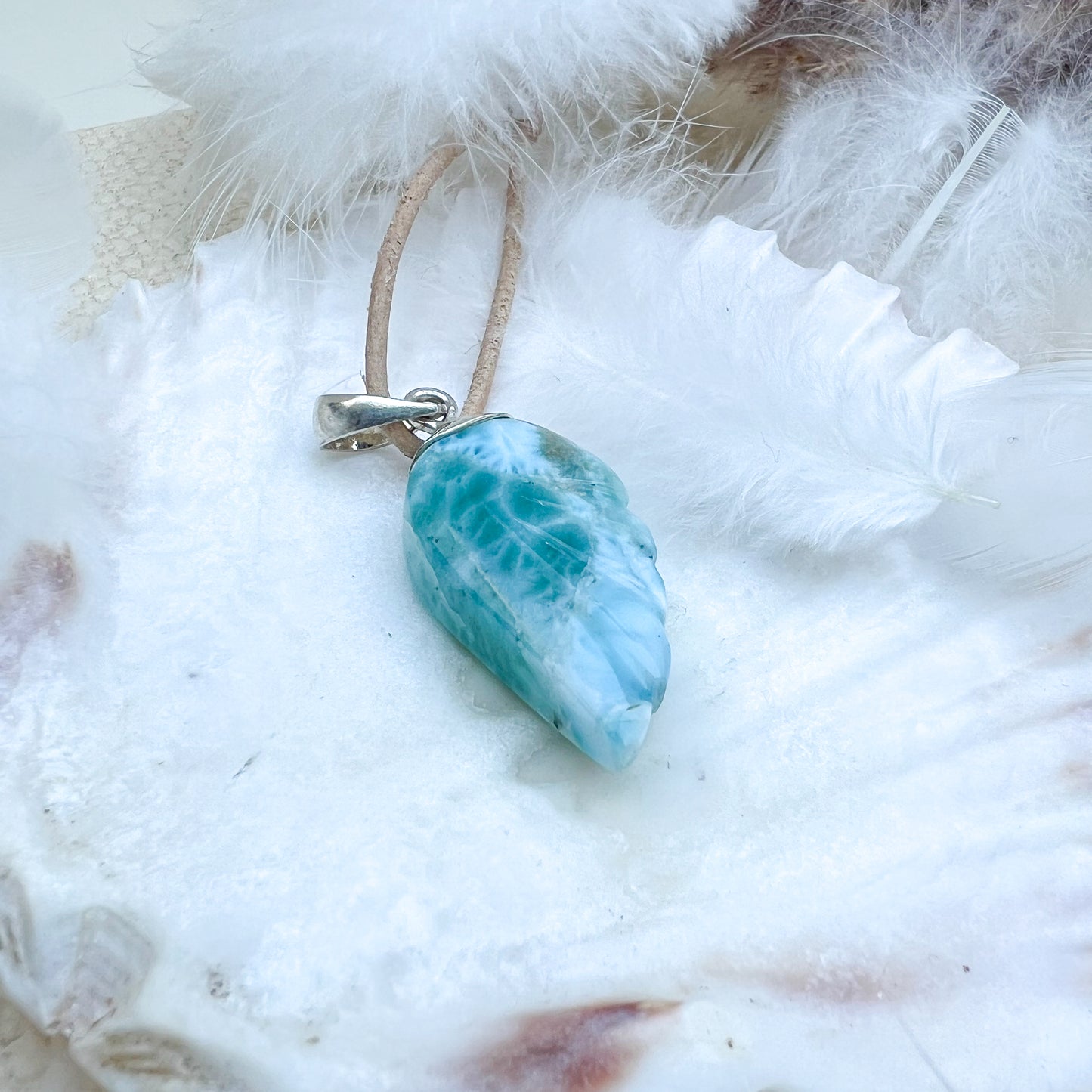 
                  
                    Larimar Engelsflügel-Anhänger aus 925er Sterlingsilber, präsentiert auf einer weißen Muschelschale, umgeben von Federn, mit natürlichem Lederband – ein himmlisches Schmuckstück.
                  
                