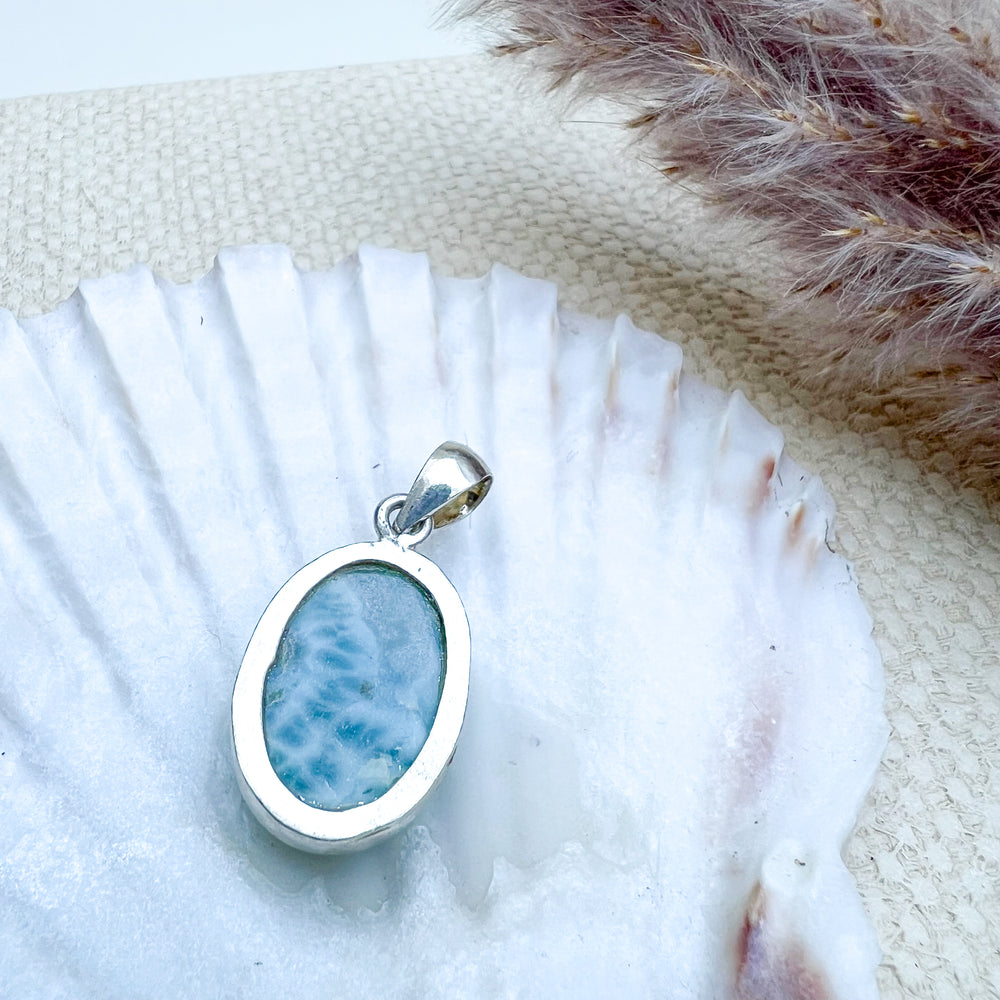 Ovaler Larimar-Anhänger aus 925er Sterlingsilber, Rückseite des Steins mit natürlicher Musterung, präsentiert auf einer weißen Muschelschale mit dekorativen Elementen.