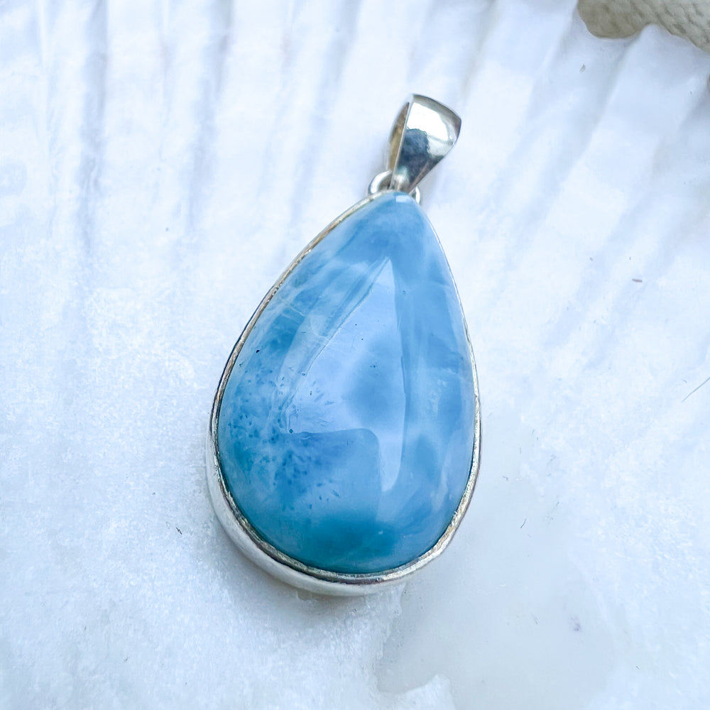 Larimar Tropfen-Anhänger aus 925er Sterlingsilber, präsentiert auf einer weißen Muschelschale, mit einer glatten, glänzenden Oberfläche und lebhaften Blau-Tönen.
