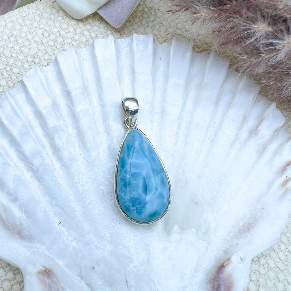 Larimar Tropfen-Anhänger aus 925er Sterlingsilber, präsentiert auf einer weißen Muschelschale, mit einer glatten, glänzenden Oberfläche und lebhaften Blau-Tönen.