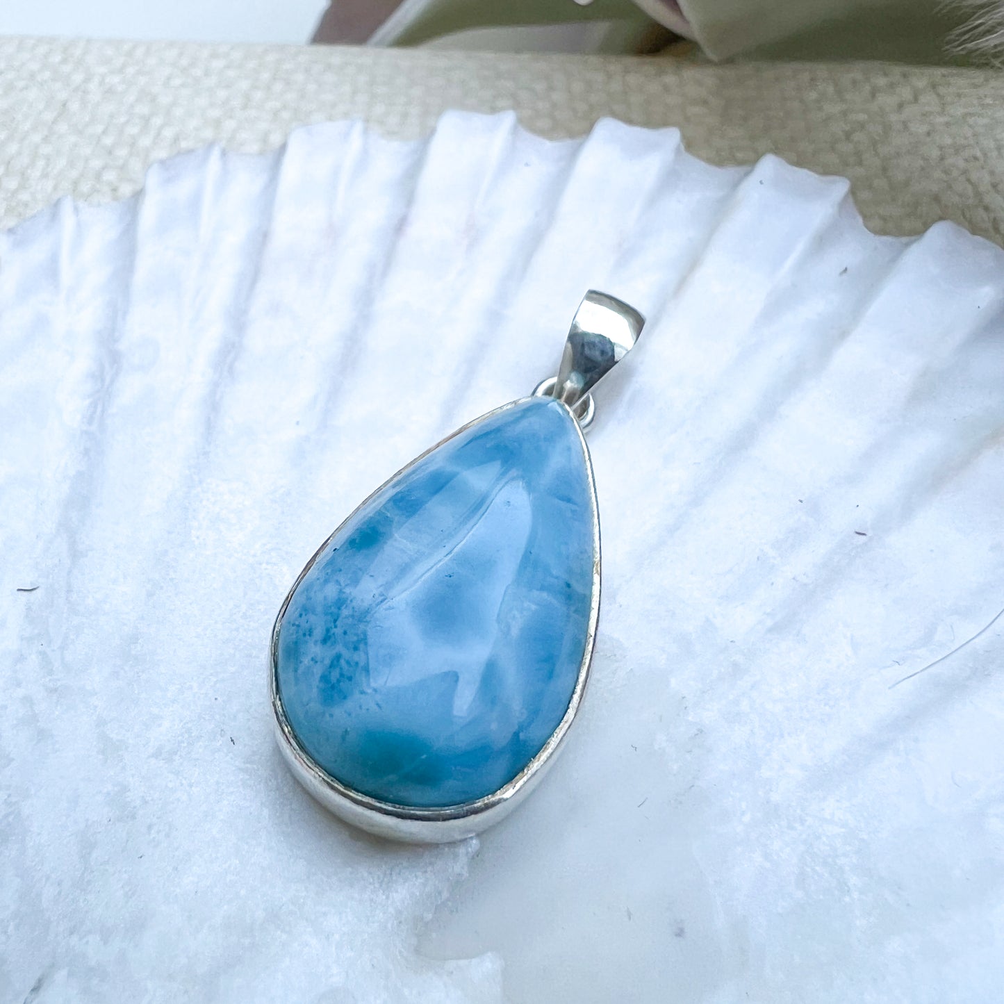 Larimar Tropfen-Anhänger aus 925er Sterlingsilber, präsentiert auf einer weißen Muschelschale, mit einer glatten, glänzenden Oberfläche und lebhaften Blau-Tönen.