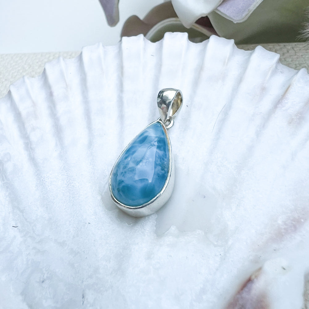 Larimar Tropfen-Anhänger aus 925er Sterlingsilber, präsentiert auf einer weißen Muschelschale, mit einer glatten, glänzenden Oberfläche und lebhaften Blau-Tönen