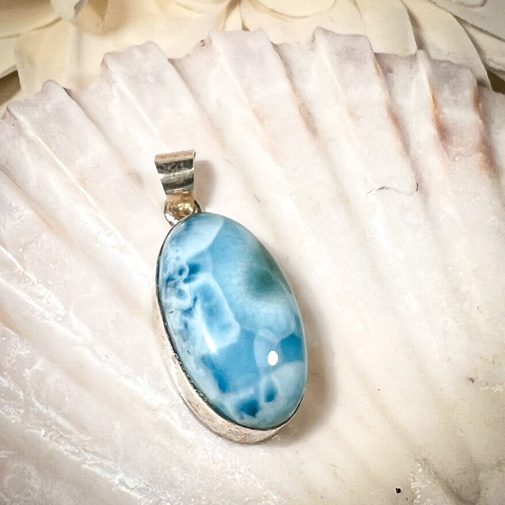 Ovaler Larimar-Kettenanhänger in 925er Sterlingsilber, auf einer Muschelschale präsentiert. Der blau-weiße Edelstein besticht durch natürliche Farbverläufe und erinnert an das karibische Meer. Einzigartiges Schmuckstück für Damen.