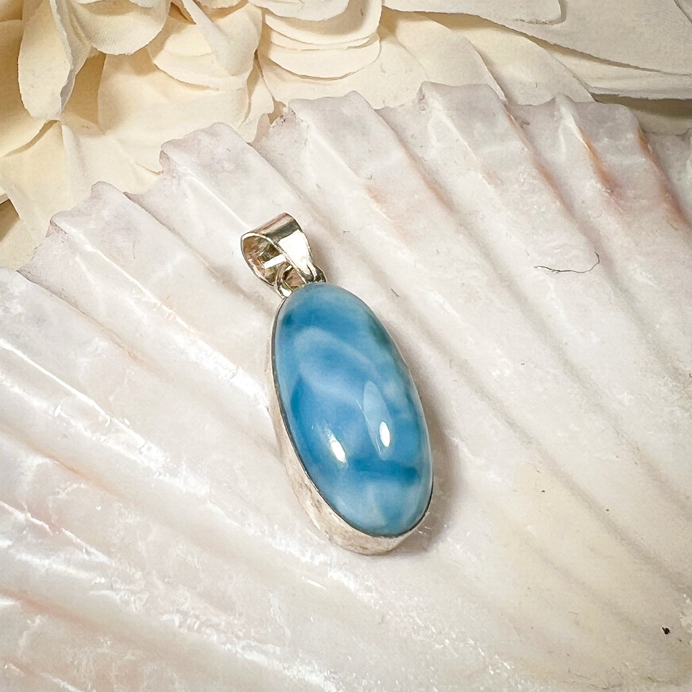 Ovaler Larimar-Kettenanhänger in 925er Sterlingsilber, auf einer Muschelschale präsentiert. Der blau-weiße Edelstein besticht durch natürliche Farbverläufe und erinnert an das karibische Meer. Einzigartiges Schmuckstück für Damen.