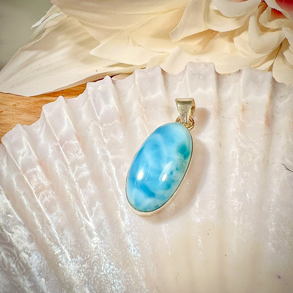 Ovaler Larimar-Kettenanhänger in 925er Sterlingsilber, auf einer Muschelschale präsentiert. Der blau-weiße Edelstein besticht durch natürliche Farbverläufe und erinnert an das karibische Meer. Einzigartiges Schmuckstück für Damen.