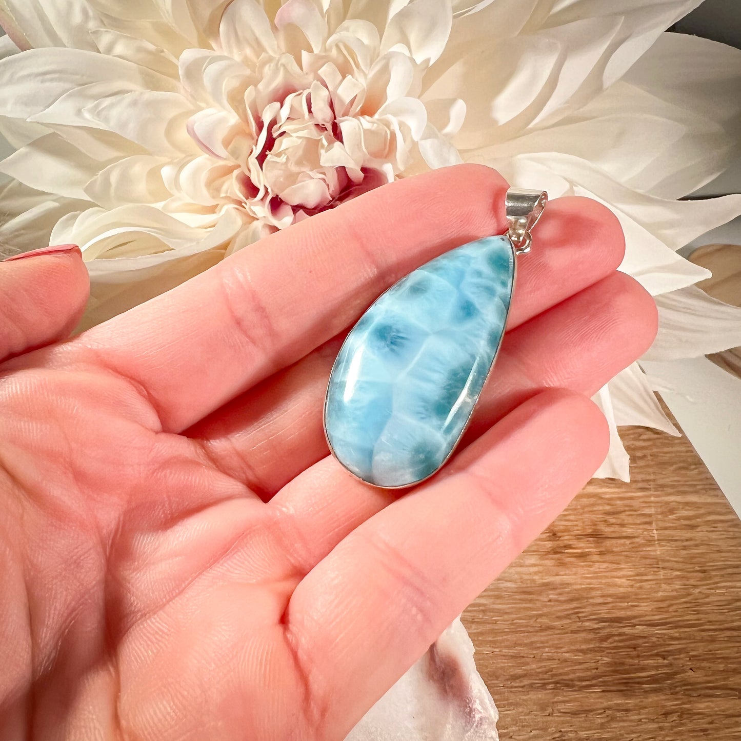 
                  
                    Hand hält einen eleganten Larimar-Anhänger in Tropfenform, gefasst in hochwertigem Silber. Der blaue Edelstein zeigt eine natürliche Maserung und wird vor einer weißen Blume präsentiert. Dieser besondere Larimar-Anhänger eignet sich perfekt als stilvolles Schmuckstück oder spirituelles Accessoire.
                  
                