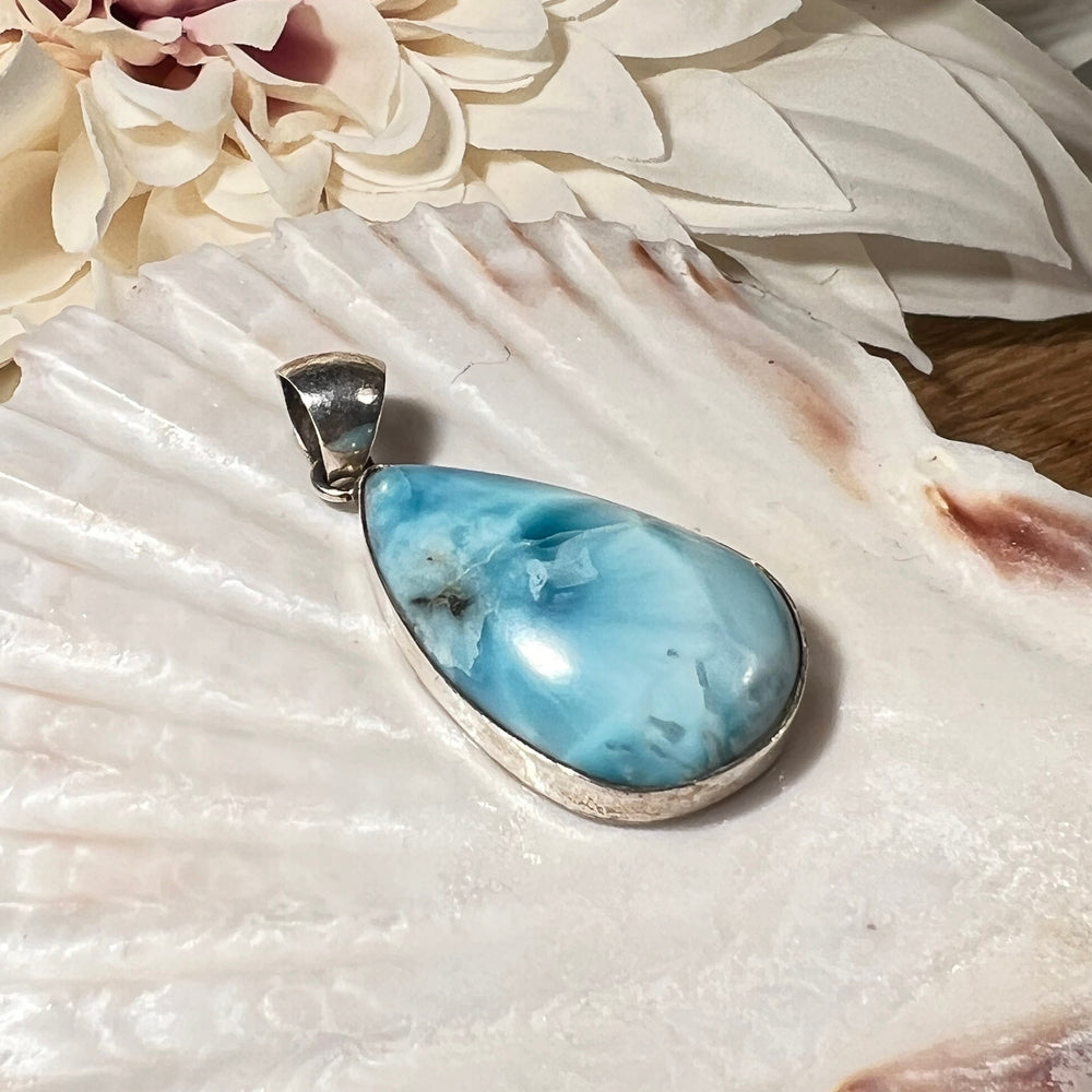 Hochwertiger Larimar-Anhänger in Tropfenform, gefasst in glänzendem Silber, präsentiert auf einer Muschelschale. Der himmelblaue Larimar-Edelstein mit natürlichen Farbverläufen und Maserungen verleiht dem Schmuckstück eine besondere Eleganz. Idealer Edelsteinanhänger für Damen, der maritimes Flair und eine spirituelle Note vereint – perfekt als stilvolles Accessoire oder besonderes Geschenk.