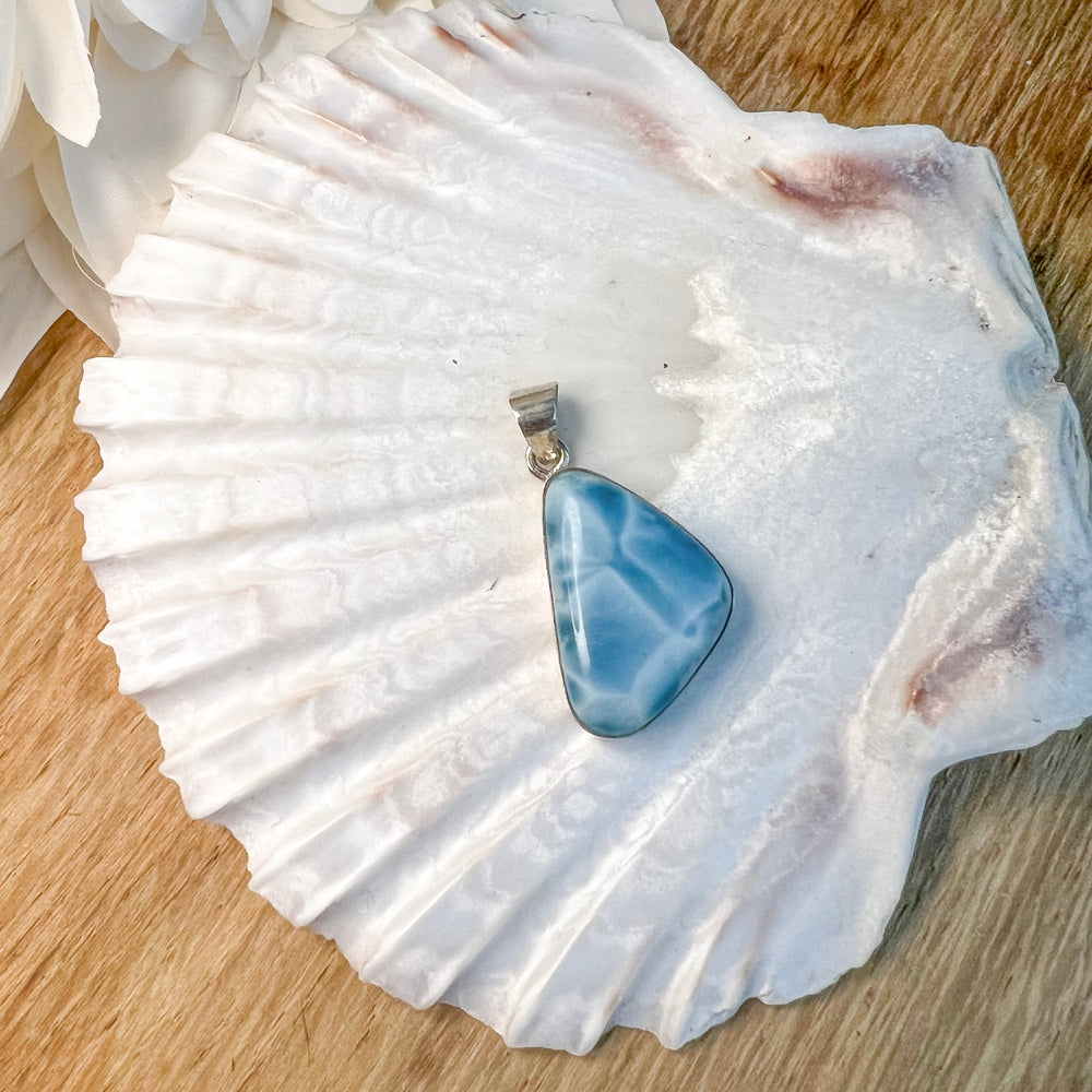 Dreieckiger Larimar-Kettenanhänger in 925er Sterlingsilber auf Muschelschale. Der Edelstein zeigt intensive Blautöne mit natürlichen weißen Adern, inspiriert vom karibischen Meer. Einzigartiges Schmuckstück und ideales Geschenk.