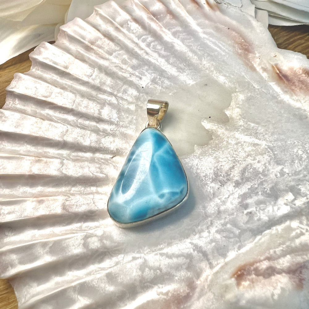 Dreieckiger Larimar-Kettenanhänger in 925er Sterlingsilber auf Muschelschale. Der Edelstein zeigt intensive Blautöne mit natürlichen weißen Adern, inspiriert vom karibischen Meer. Einzigartiges Schmuckstück und ideales Geschenk.