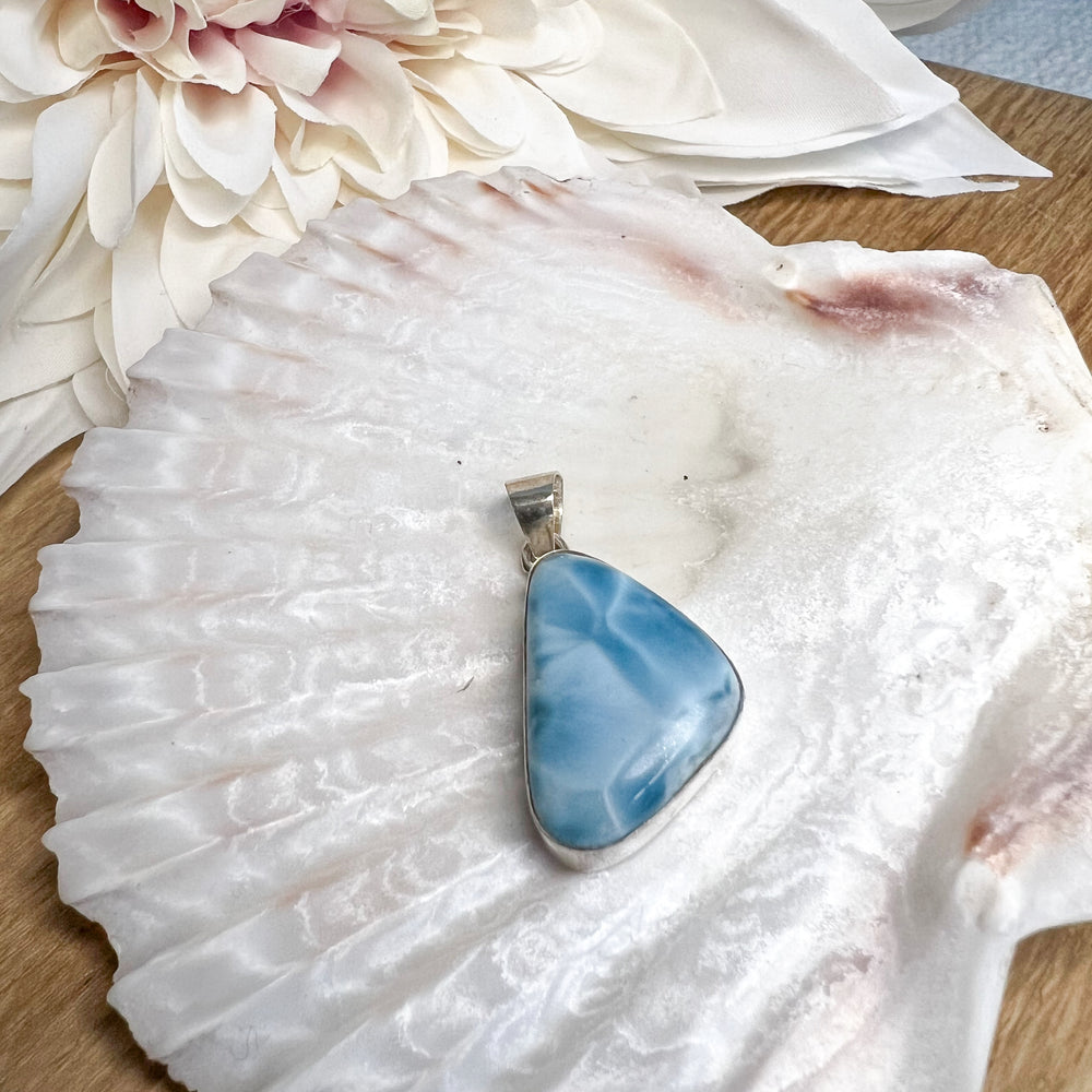 Dreieckiger Larimar-Kettenanhänger in 925er Sterlingsilber auf Muschelschale. Der Edelstein zeigt intensive Blautöne mit natürlichen weißen Adern, inspiriert vom karibischen Meer. Einzigartiges Schmuckstück und ideales Geschenk.