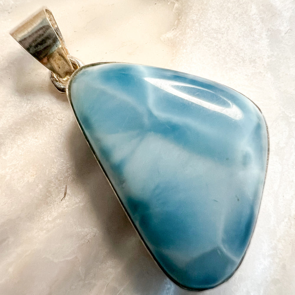 Dreieckiger Larimar-Kettenanhänger in 925er Sterlingsilber auf Muschelschale. Der Edelstein zeigt intensive Blautöne mit natürlichen weißen Adern, inspiriert vom karibischen Meer. Einzigartiges Schmuckstück und ideales Geschenk.