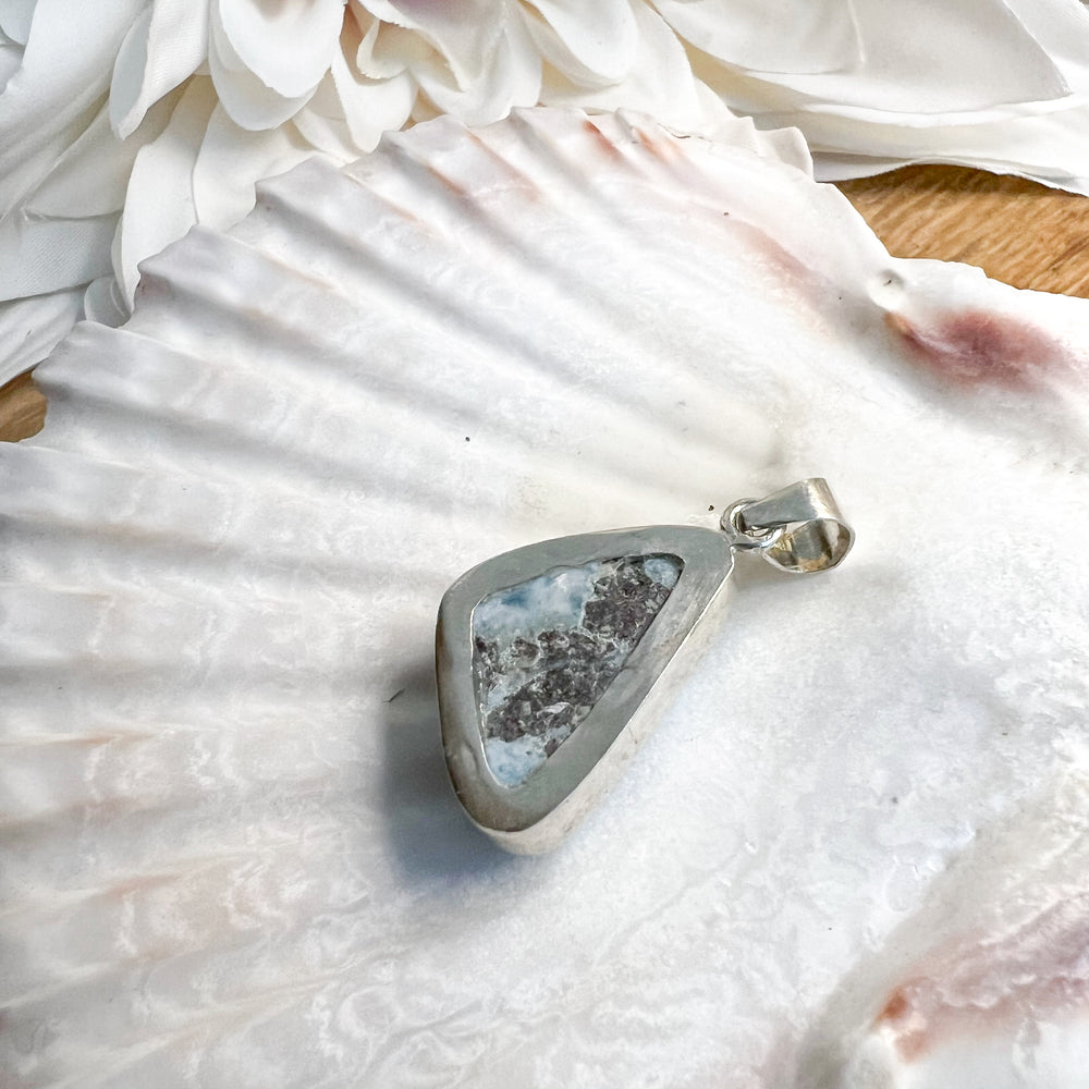 Dreieckiger Larimar-Kettenanhänger in 925er Sterlingsilber auf Muschelschale. Der Edelstein zeigt intensive Blautöne mit natürlichen weißen Adern, inspiriert vom karibischen Meer. Einzigartiges Schmuckstück und ideales Geschenk.