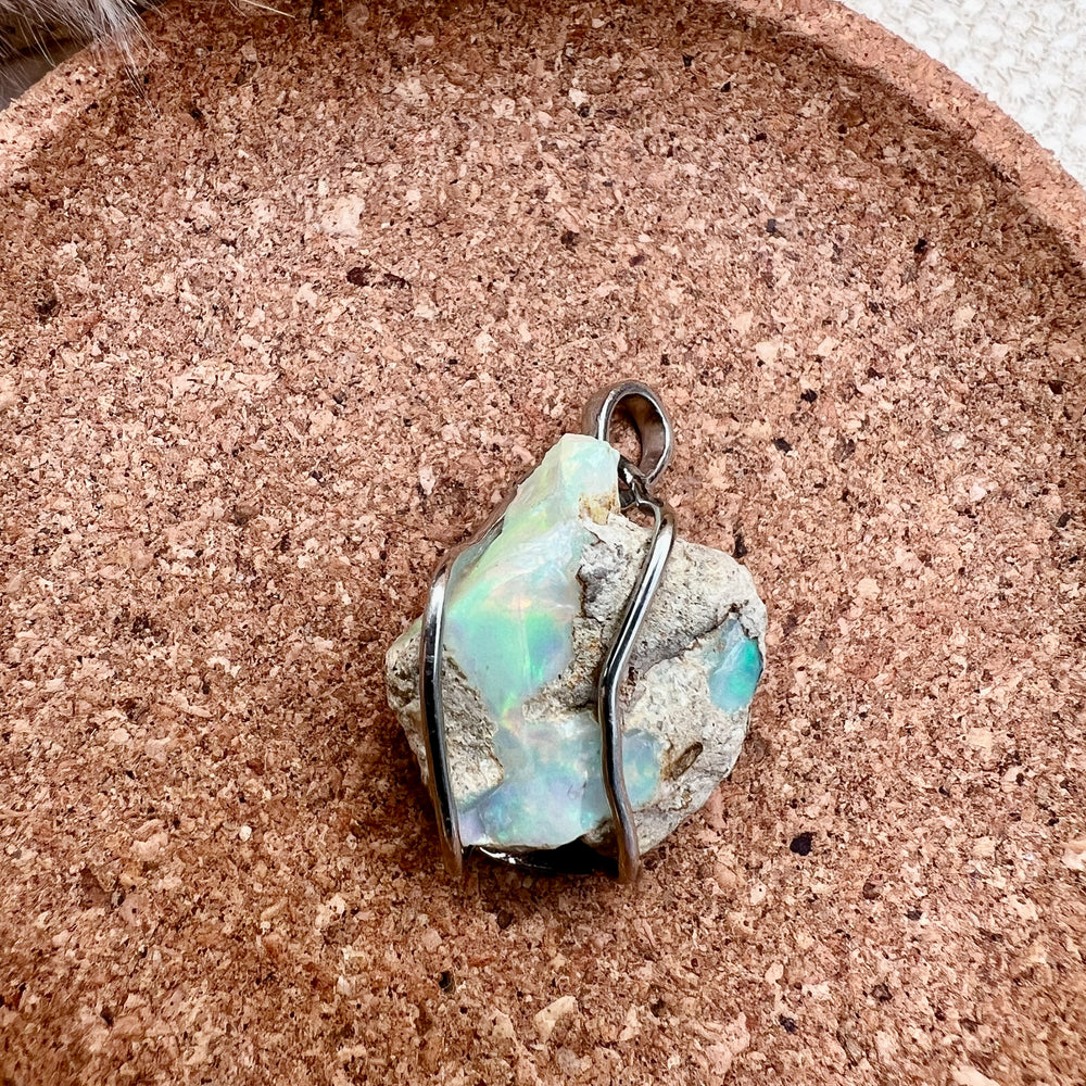 Ein natürlicher Opal-Rohstein mit schimmernden Grüntönen und rauen Strukturen, gefasst in eine elegante Silberfassung als Kettenanhänger. Der Schmuck liegt auf einer runden Unterlage aus Kork, während im Hintergrund unscharfe, getrocknete Gräser eine warme und natürliche Atmosphäre schaffen.