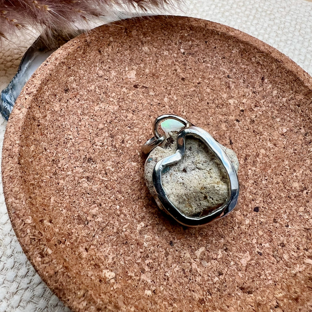 Ein natürlicher Opal-Rohstein mit schimmernden Grüntönen und rauen Strukturen, gefasst in eine elegante Silberfassung als Kettenanhänger. Der Schmuck liegt auf einer runden Unterlage aus Kork, während im Hintergrund unscharfe, getrocknete Gräser eine warme und natürliche Atmosphäre schaffen.