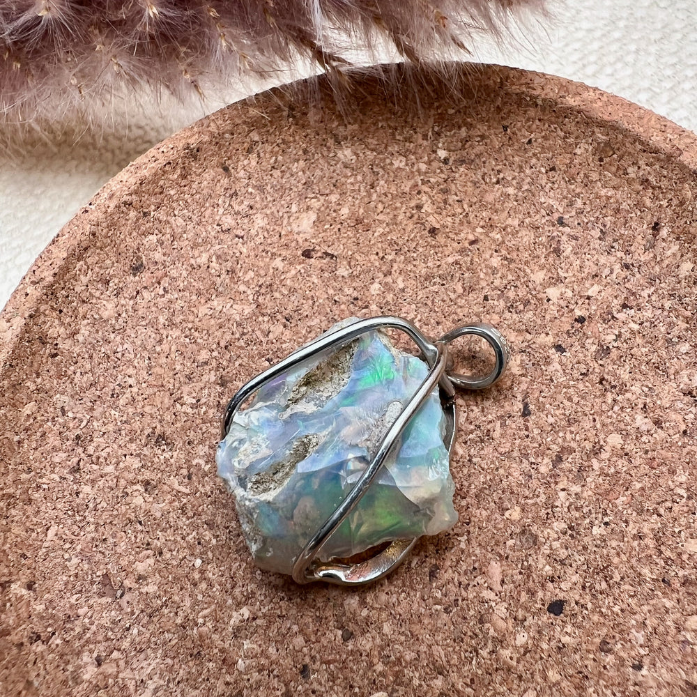 
                  
                    Ein natürlicher Opal-Rohstein mit schimmernden Grüntönen und rauen Strukturen, gefasst in eine elegante Silberfassung als Kettenanhänger. Der Schmuck liegt auf einer runden Unterlage aus Kork, während im Hintergrund unscharfe, getrocknete Gräser eine warme und natürliche Atmosphäre schaffen.
                  
                