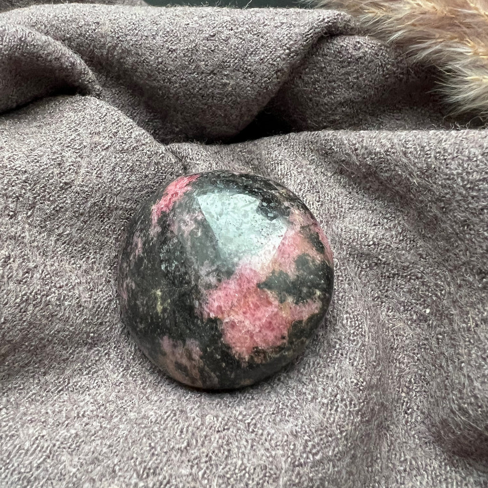 Ein polierter Rhodonit Handschmeichler in intensiven Rosa- und Rottönen mit natürlichen schwarzen und goldenen Einschlüssen, präsentiert auf einem weichen grauen Stoffhintergrund.